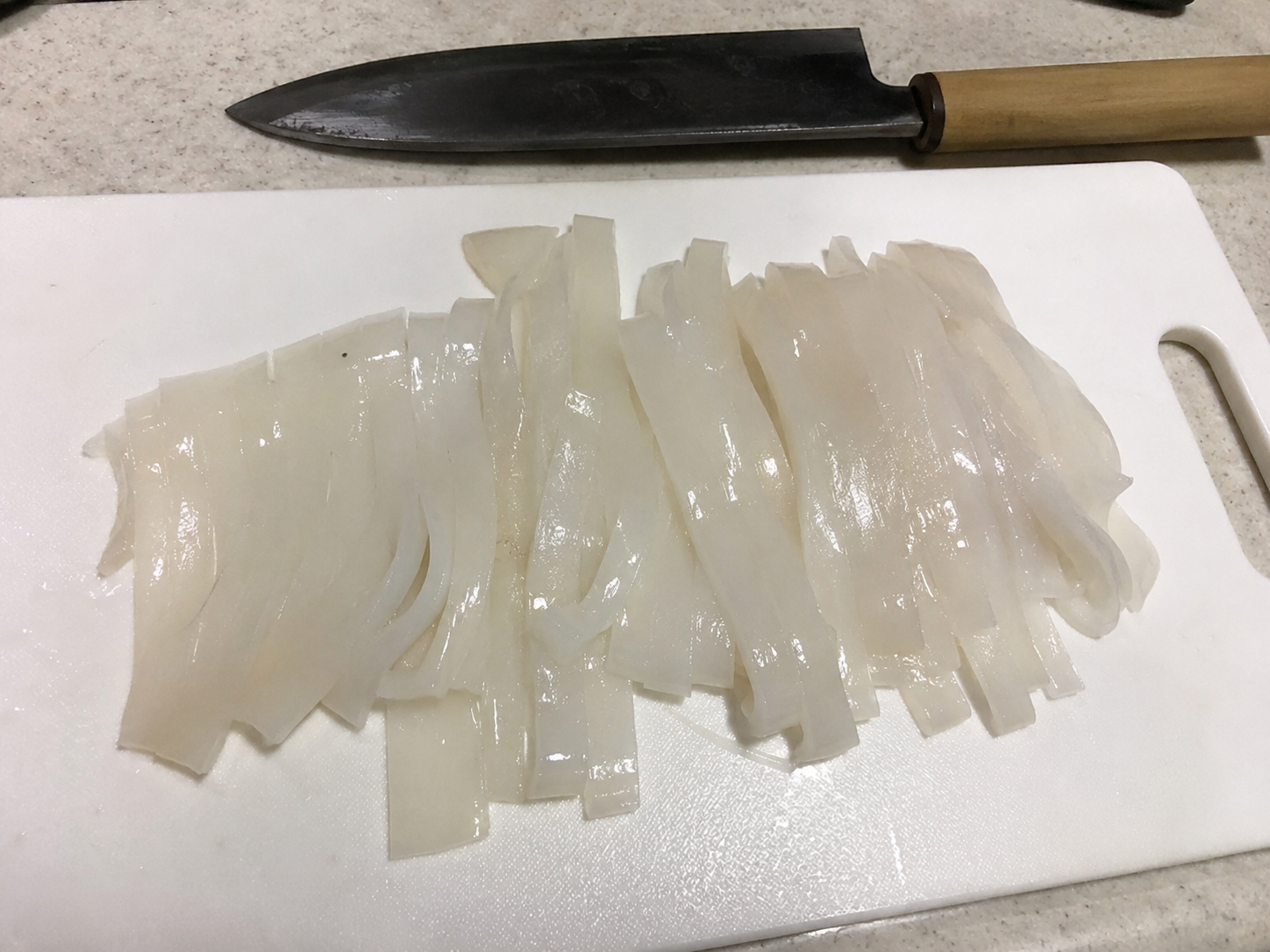 家庭のキッチンで白いまな板の上に切られたイカそうめんが並び、奥に包丁が置かれているリアルな自炊風写真。盛り付けは簡素で、生活感のある雰囲気。